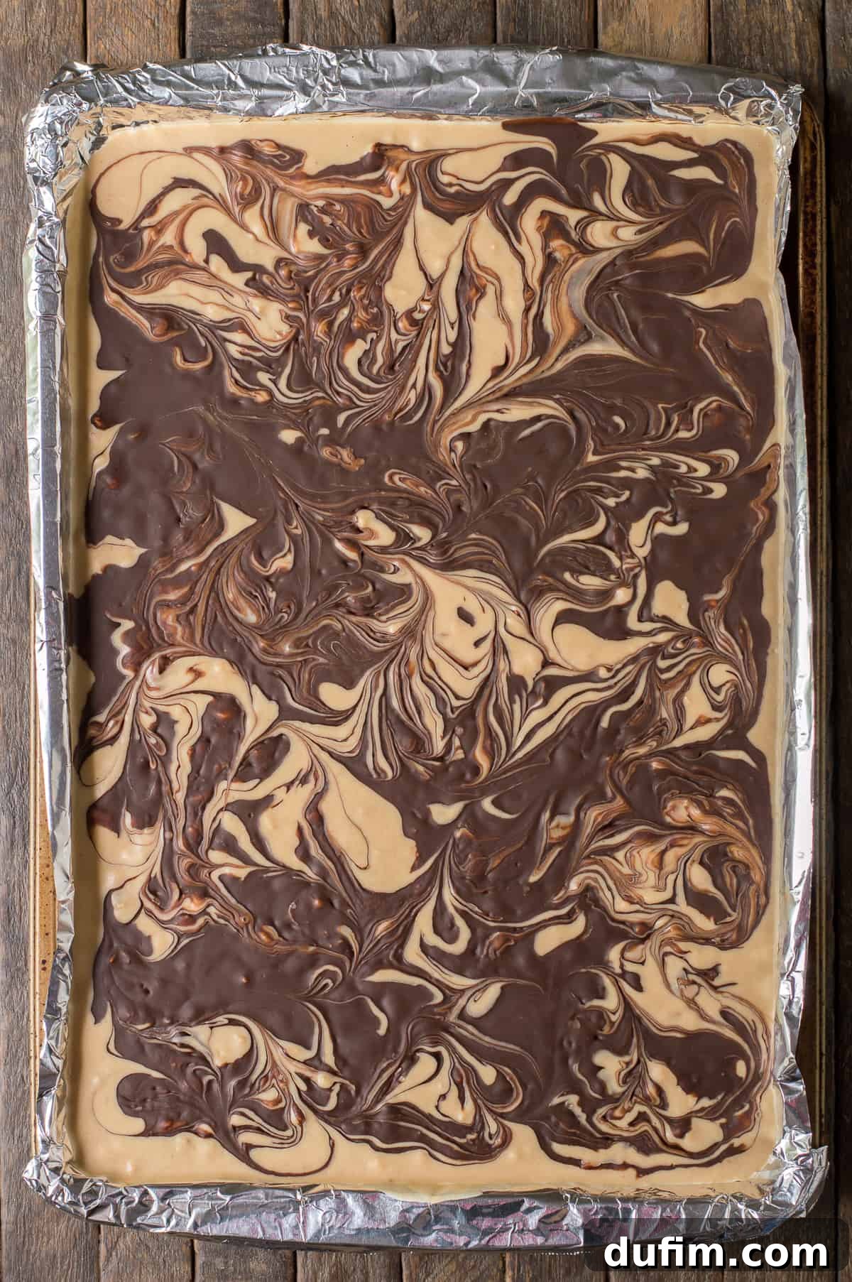Tiger butter mixture setting in a jelly roll pan lined with aluminum foil, showing the initial swirl pattern.