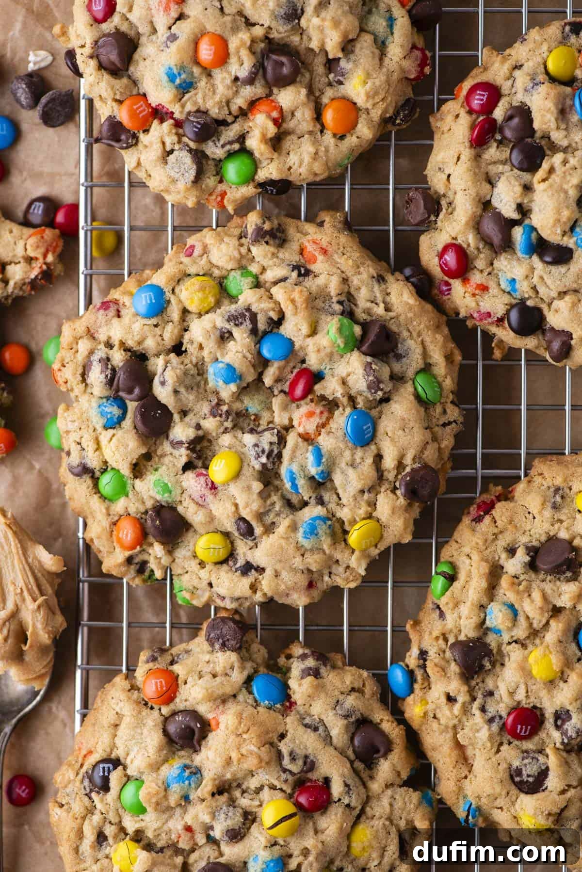 Large, vibrant monster cookies filled with colorful candy-coated chocolates and melty chocolate chips cooling on a wire rack. Nearby, a spoonful of creamy peanut butter and scattered cookie crumbs hint at the delicious ingredients.