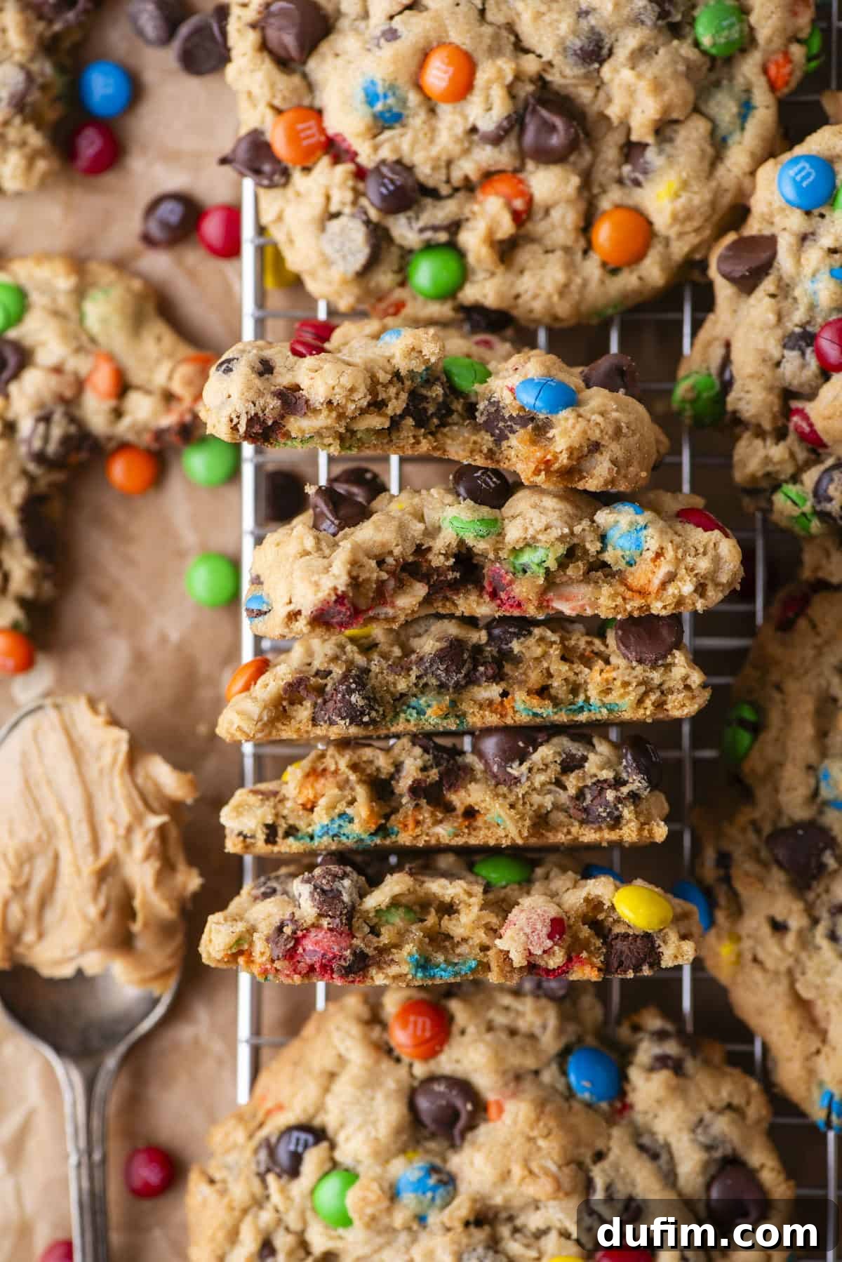 A stack of thick, wonderfully chewy monster cookies, generously filled with colorful chocolate candies and rich chocolate chips, rests invitingly on a cooling rack. One cookie is broken in half, revealing its incredibly soft and tender texture inside. Nearby, a spoonful of creamy peanut butter hints at a key ingredient.