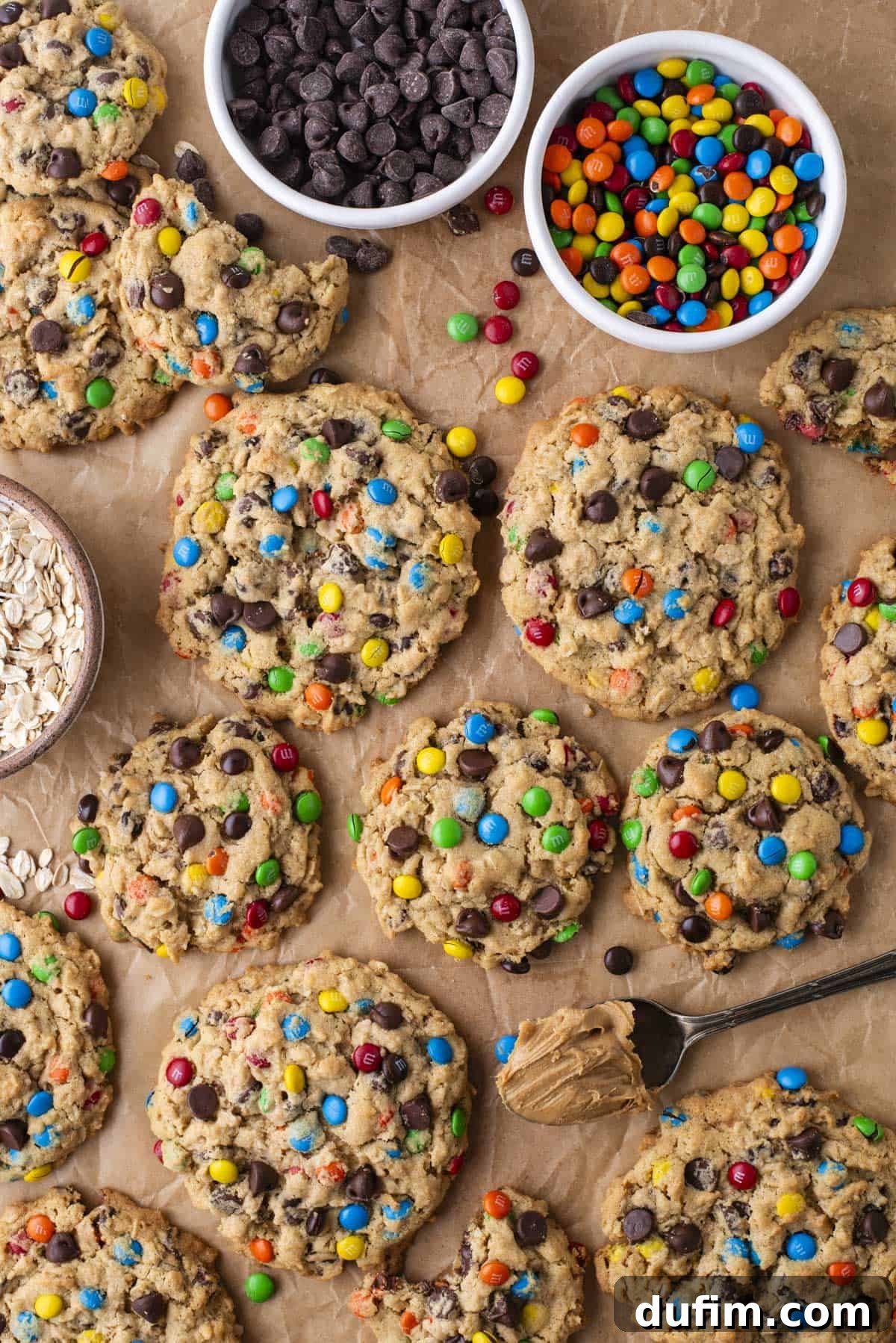 A batch of large, perfectly baked monster cookies with vibrant chocolate candies and chocolate chips rests on parchment paper on a baking sheet. Nearby are inviting bowls of chocolate chips, colorful M&M candies, old-fashioned oats, and a spoonful of creamy peanut butter, highlighting the core ingredients that make these cookies so irresistible.
