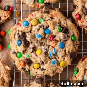A large, chunky cookie filled with colorful candy-coated chocolates and chocolate chips sits on a cooling rack, surrounded by other similar cookies.