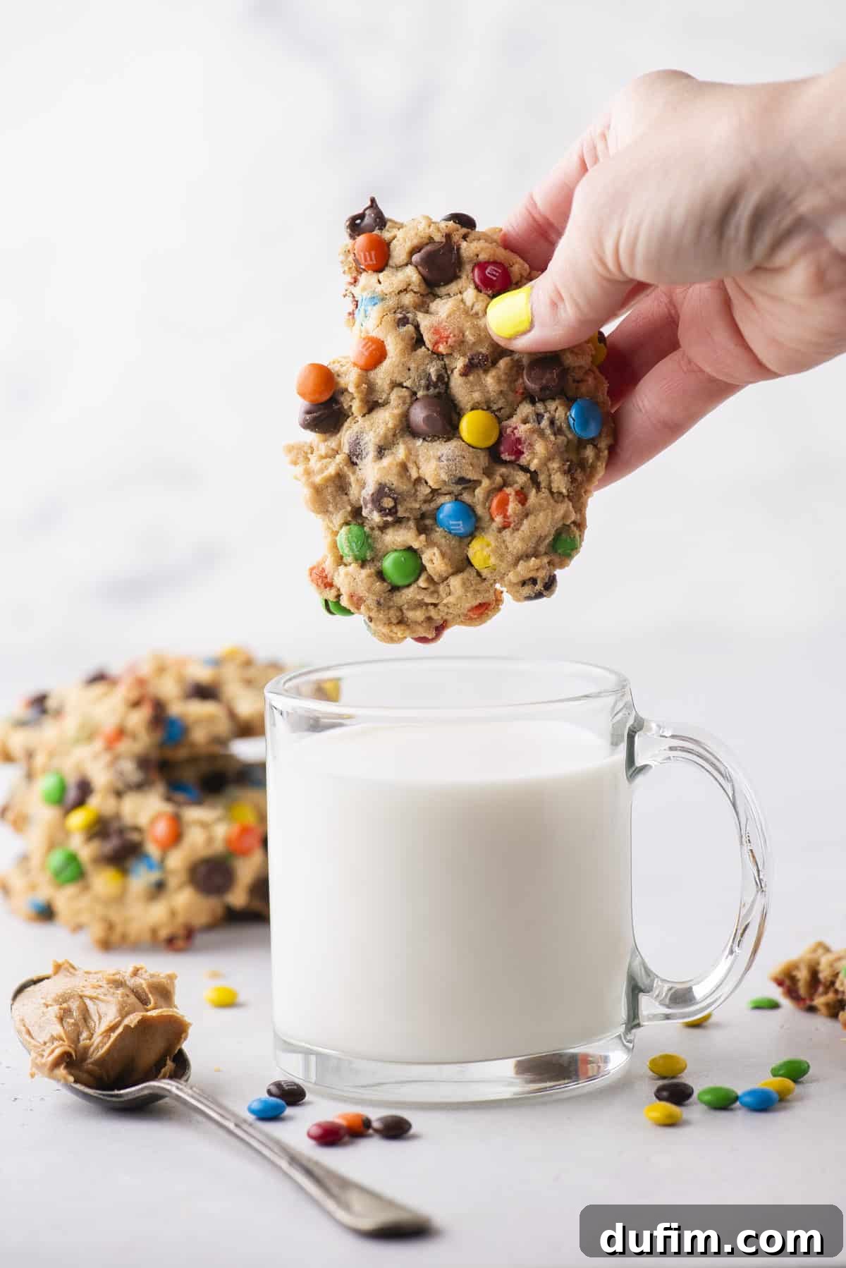 A hand dips a vibrant, candy-studded monster cookie into a clear glass mug of milk, creating a classic and inviting scene. More freshly baked cookies, a spoon with creamy peanut butter, and scattered colorful candies adorn the pristine white surface nearby, hinting at the key flavors and ingredients within these delightful treats.