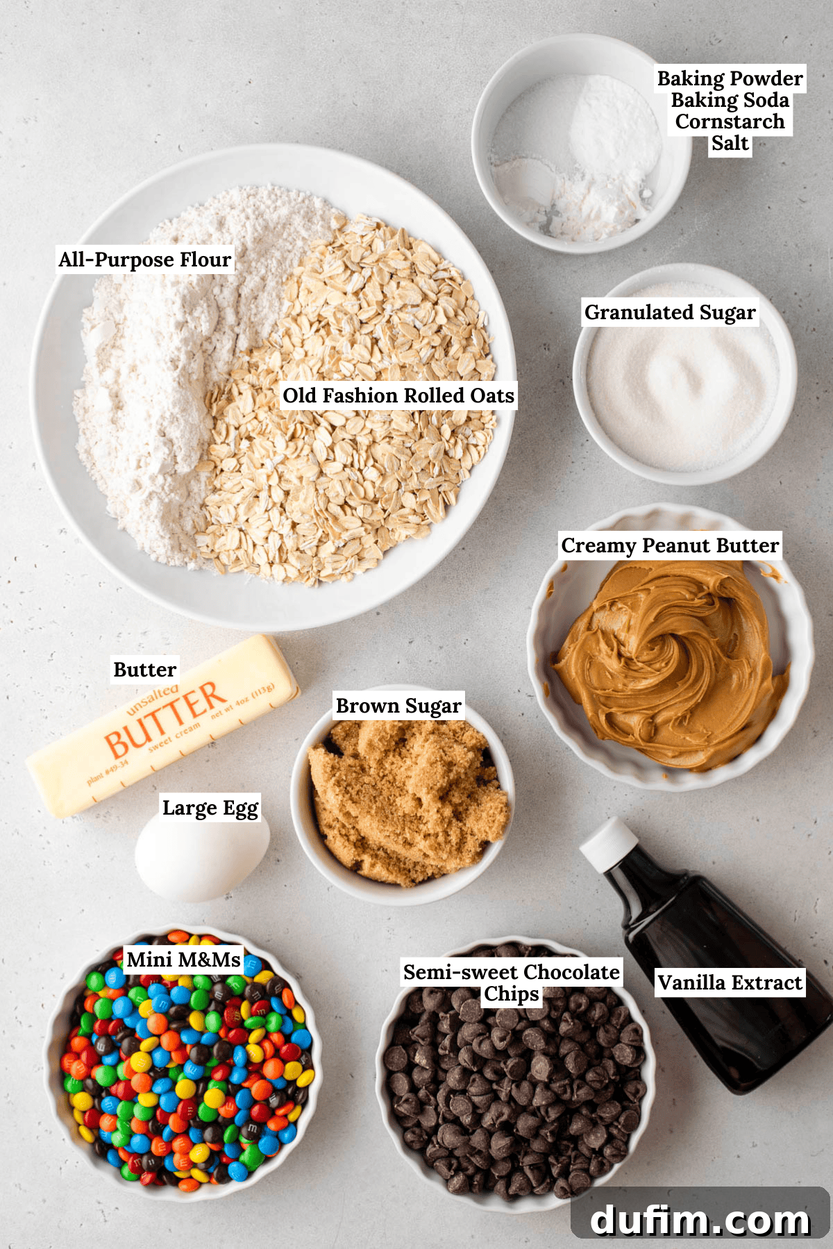 A clear, top-down photograph showcasing all the essential ingredients for making these monster cookies: neatly arranged piles of flour, old-fashioned rolled oats, granulated sugar, light brown sugar, softened butter, a large egg, creamy peanut butter, baking powder, baking soda, cornstarch, salt, vanilla extract, rich chocolate chips, and vibrant mini M&Ms.