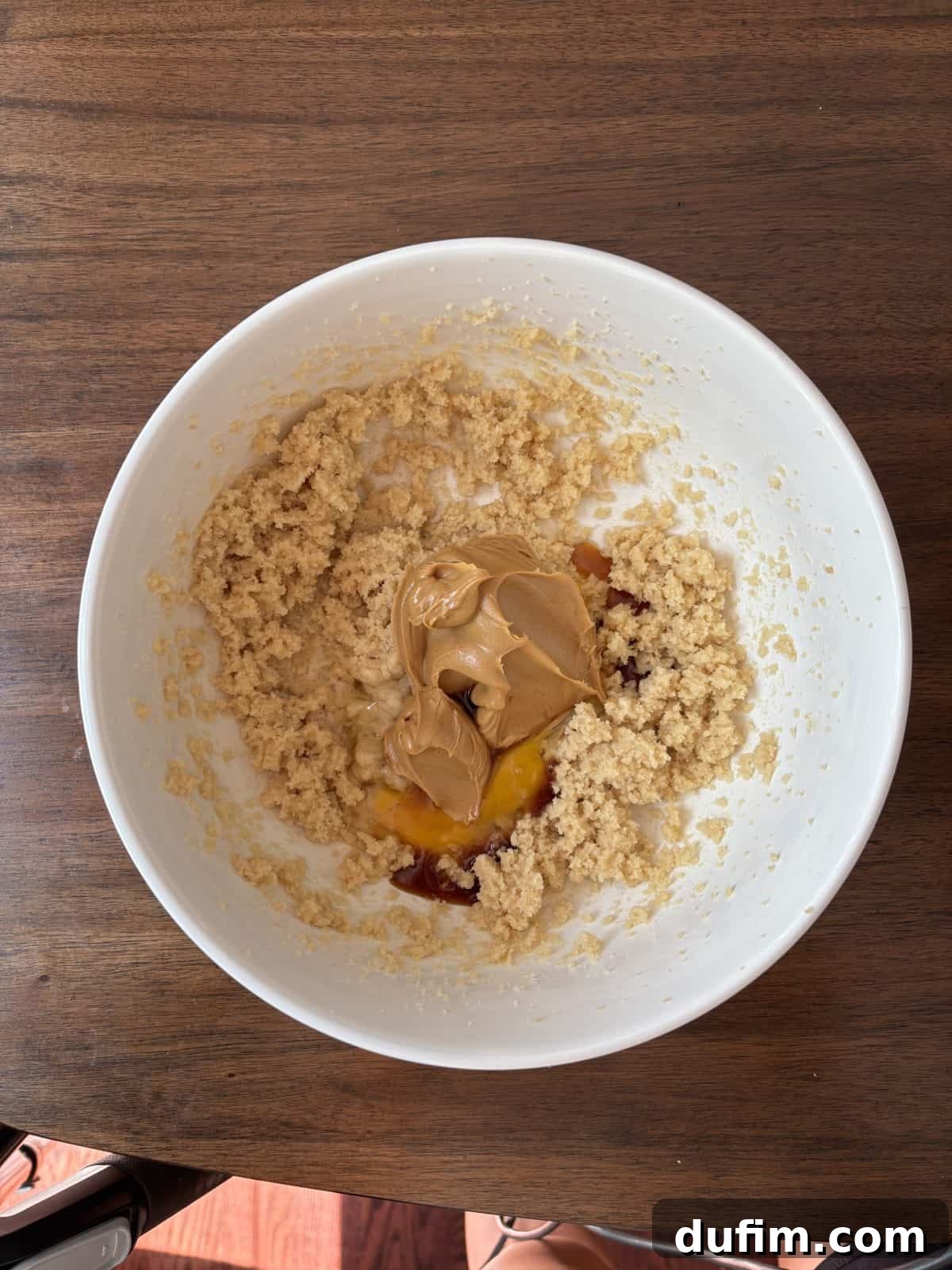 A white bowl on a rustic wooden table contains a perfectly creamed mixture of sugar and butter, with creamy peanut butter, a whole egg, and clear vanilla extract added but not yet incorporated, ready for mixing to create delicious cookie dough.
