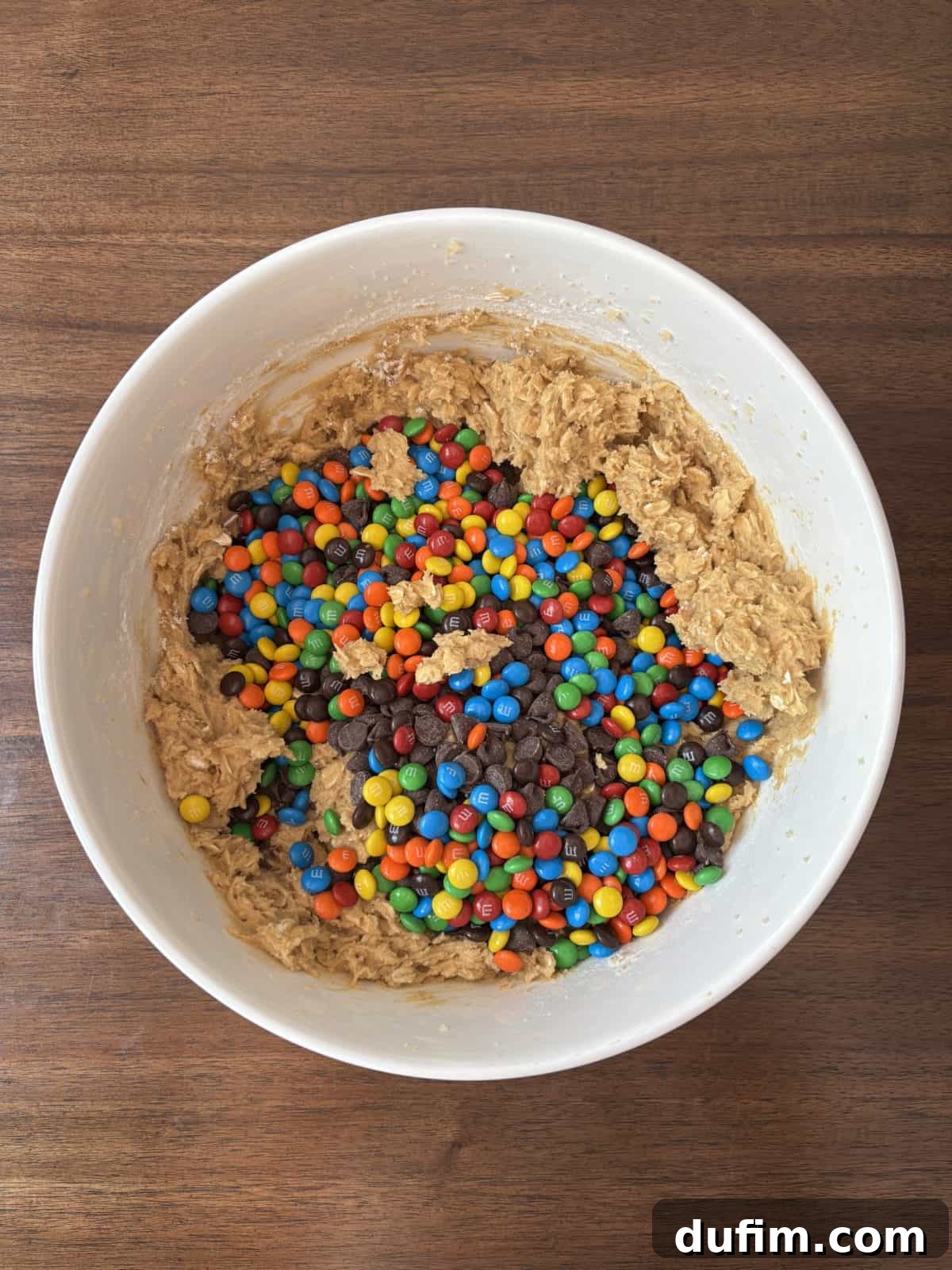 A large white mixing bowl resting on a warm wooden surface, filled with richly textured cookie dough. The dough is generously speckled with vibrant, colorful mini chocolate candies and delectable chocolate chips, promising a burst of flavor in every bite.
