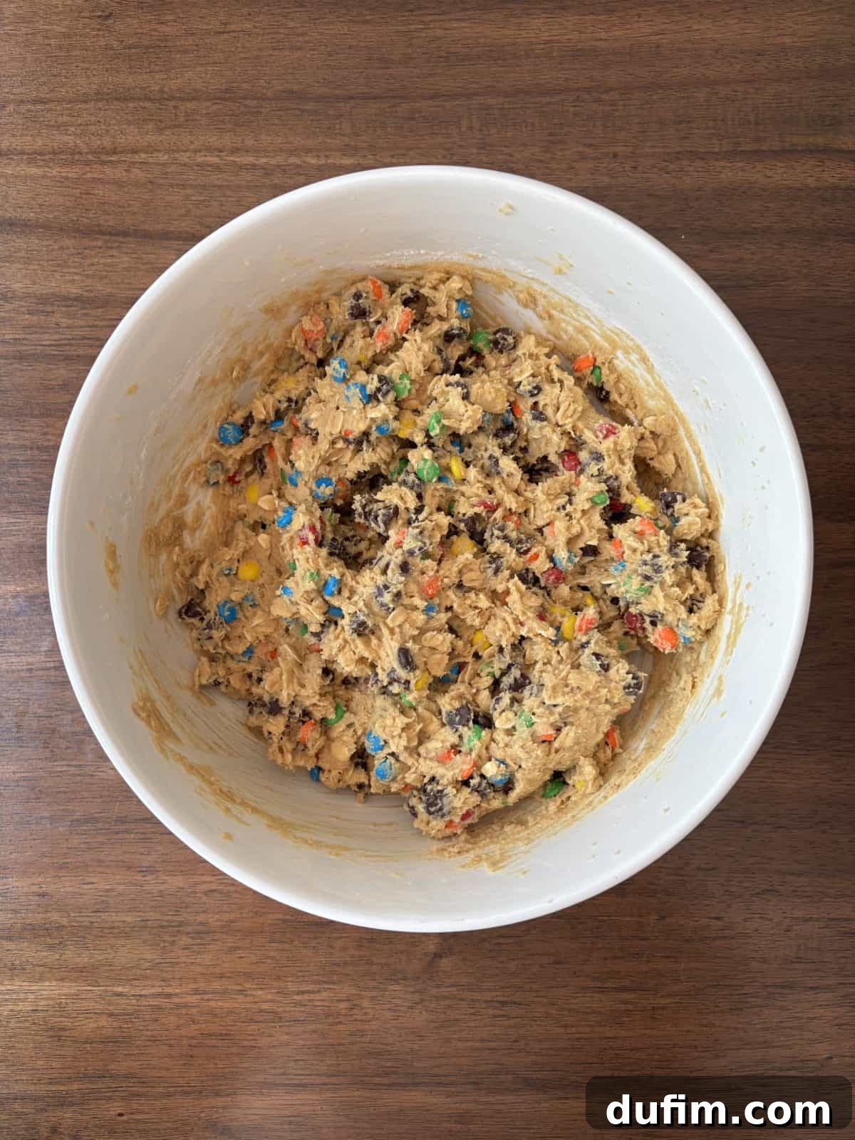 A pristine white bowl brimming with unbaked cookie dough, expertly mixed with a vibrant assortment of chocolate chips and colorful candy pieces, patiently awaiting its transformation in the oven, resting on a rustic wooden surface, ready for baking.
