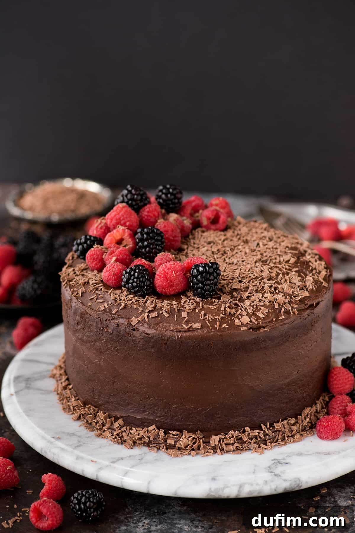 A beautifully frosted two-layer chocolate velvet cake adorned with fresh raspberries and blackberries, with delicate chocolate shavings scattered on top, presented on an elegant cake stand.