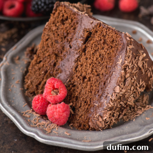 A slice of rich chocolate velvet cake with chocolate frosting and chocolate shavings on a gray plate, garnished with three fresh raspberries.