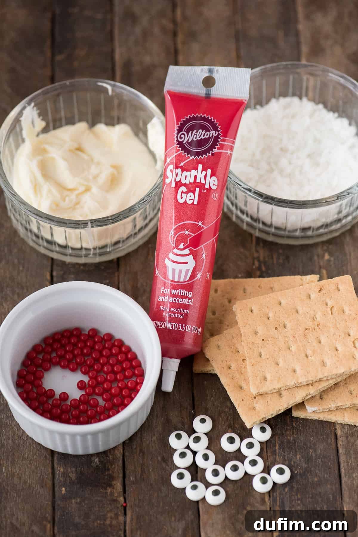 Various ingredients for Santa Graham Crackers in clear bowls, including graham crackers, frosting, shredded coconut, and red sparkle gel, ready for assembly.