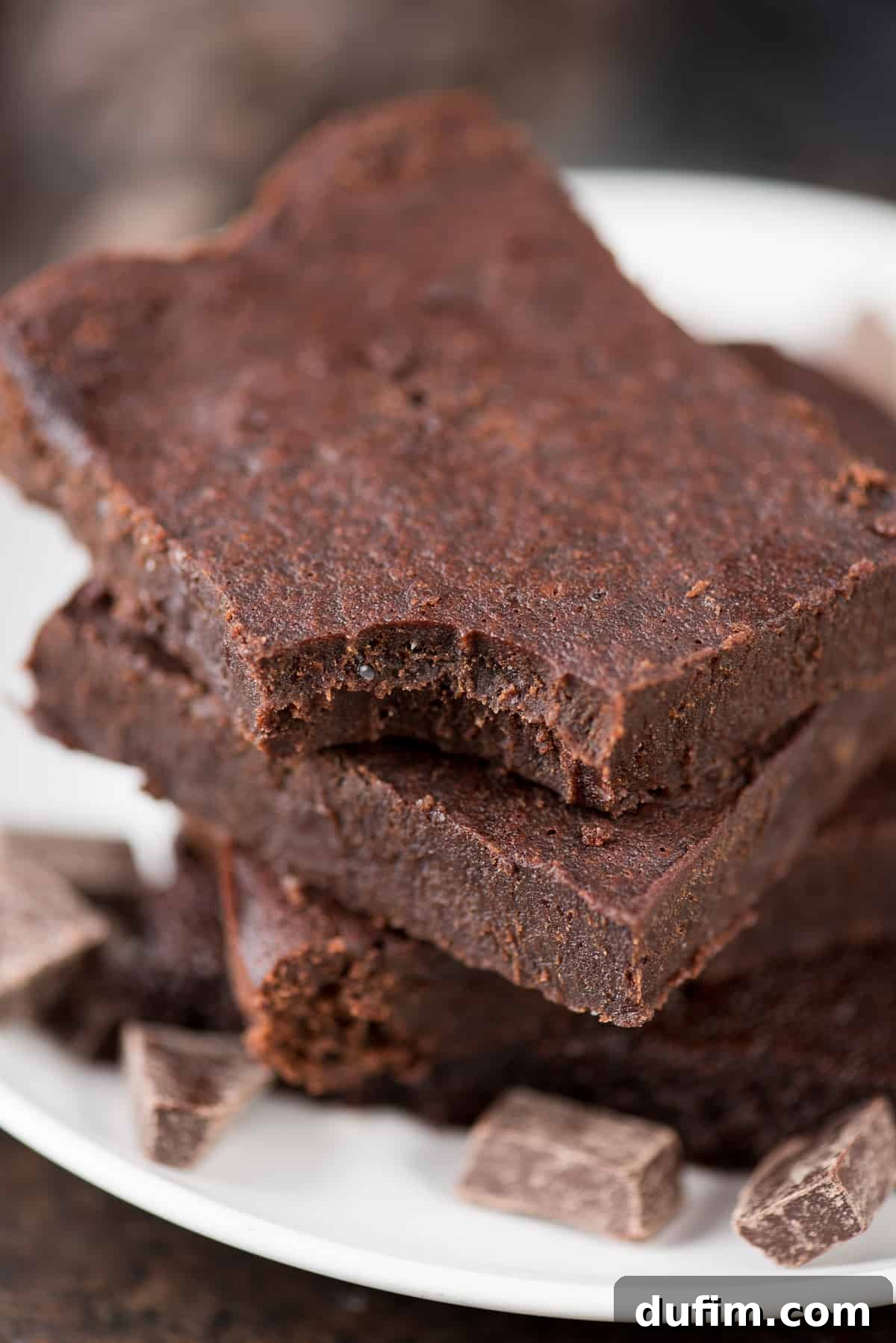 Three healthy fudgy brownies surrounded by small chocolate pieces on a white serving plate, highlighting their rich color and texture.