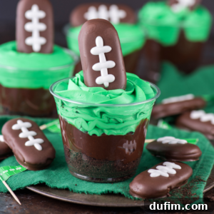 A clear cup filled with a chocolate football dirt cup, topped with green whipped cream and a football-shaped cookie decorated with white icing laces. More cups and cookies sit on a green napkin in the background.