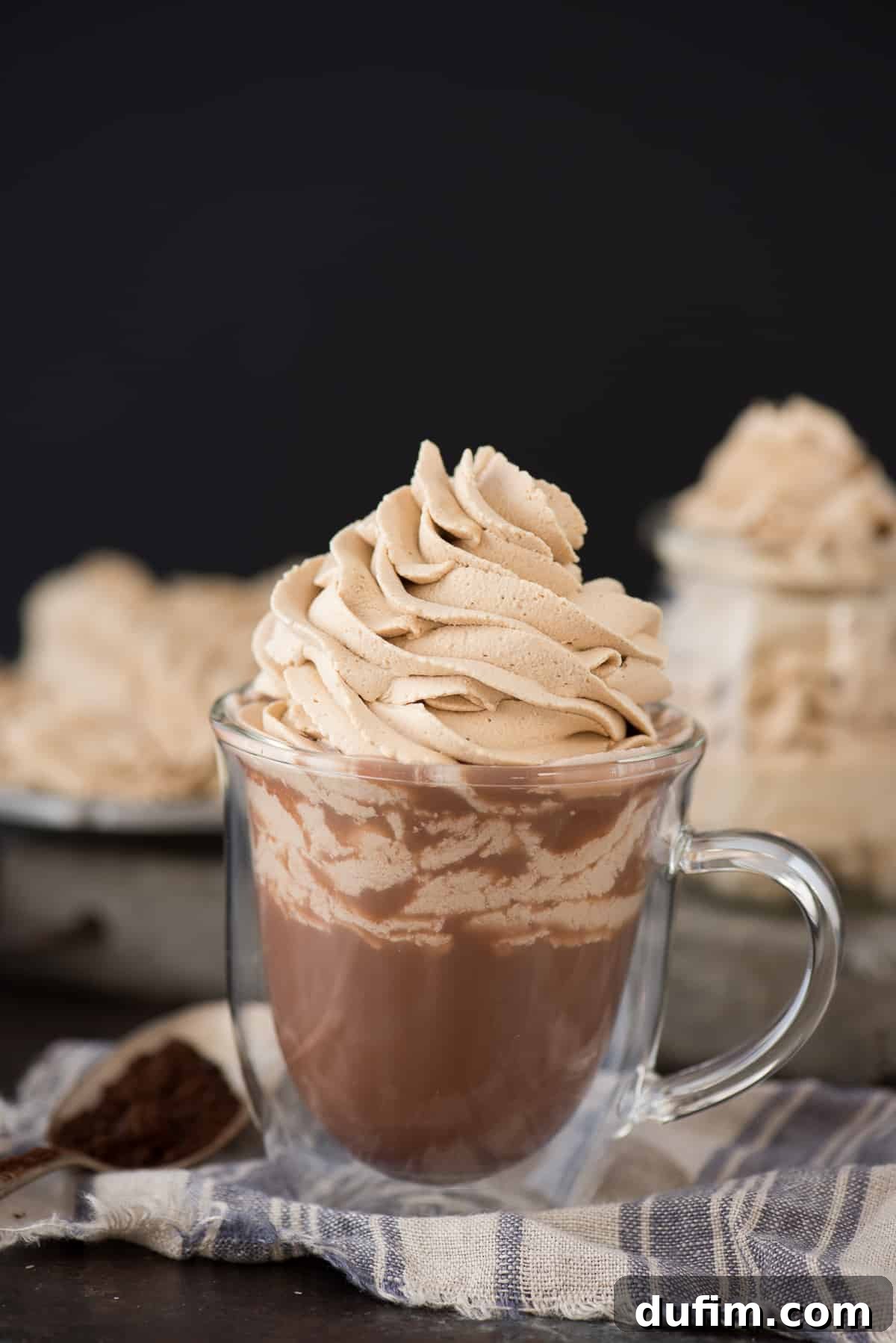 A clear glass mug of rich hot chocolate crowned with a generous, fluffy swirl of homemade coffee whipped cream, presented against a dark, inviting background.