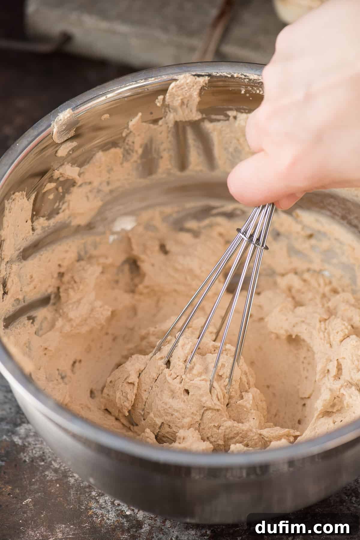 A stainless steel whisk is captured mid-motion, stirring freshly whipped coffee cream in a gleaming metal mixing bowl, highlighting its smooth and airy texture.