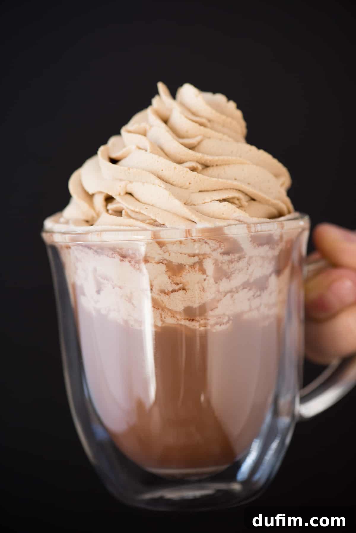 A close-up shot of a clear glass mug filled with steamy hot cocoa, generously topped with a cloud of fluffy coffee whipped cream and a delicate sprinkle of chocolate shavings.