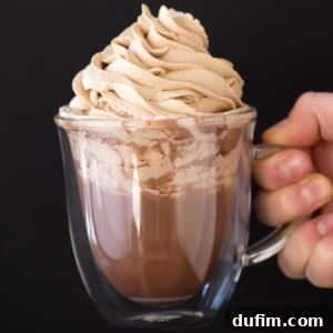 A hand holds a clear glass mug filled with hot chocolate, topped with a generous swirl of coffee whipped cream against a plain black background.