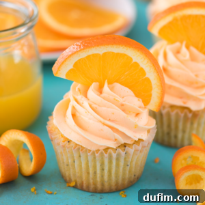 A frosted Orange Poppy Seed Cupcake adorned with a fresh orange slice, surrounded by orange peels and a jar of orange juice, on a turquoise background.