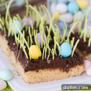 An egg hunt sugar cookie bar topped with chocolate frosting, green edible grass, and pastel candy eggs resembles a spring garden scene. The treat sits on a white plate with more candy eggs scattered nearby.