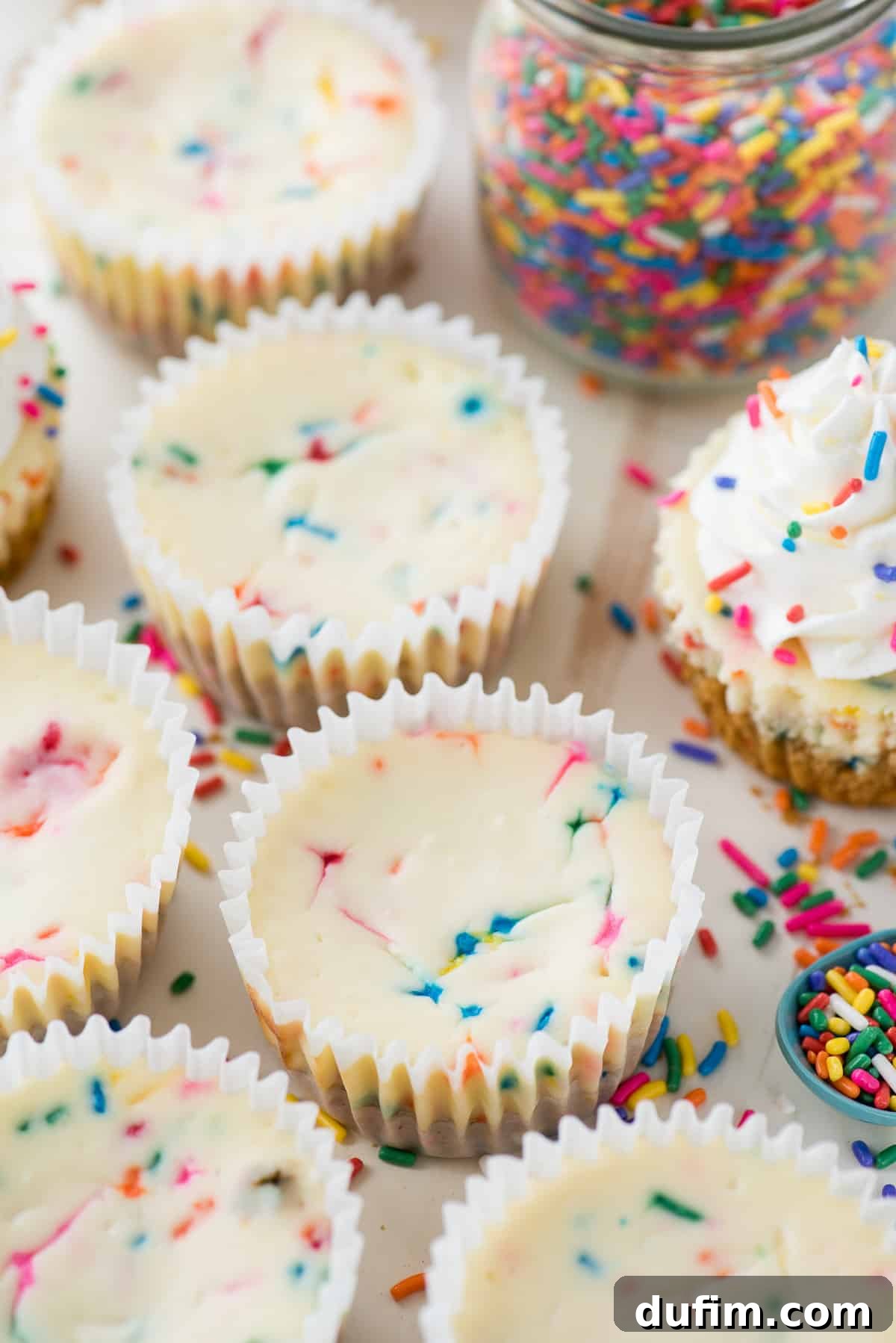 Three perfectly baked mini funfetti cheesecakes are lined up, each adorned with a scattering of colorful rainbow sprinkles on top.
