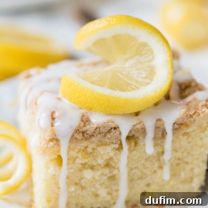 A thick slice of lemon crumb cake with a crumbly top, drizzled with white icing and garnished with two fresh lemon slices, sits on a white plate. Whole and sliced lemons are blurred in the background, enhancing the fresh lemon theme.