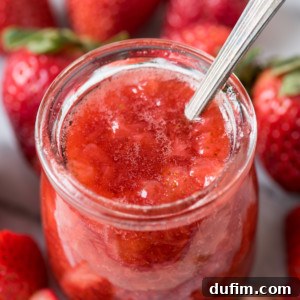A glass jar filled with chunky strawberry jam, resembling a thick strawberry sauce, sits with a spoon inside. Fresh whole and sliced strawberries are scattered around the jar on a light surface.