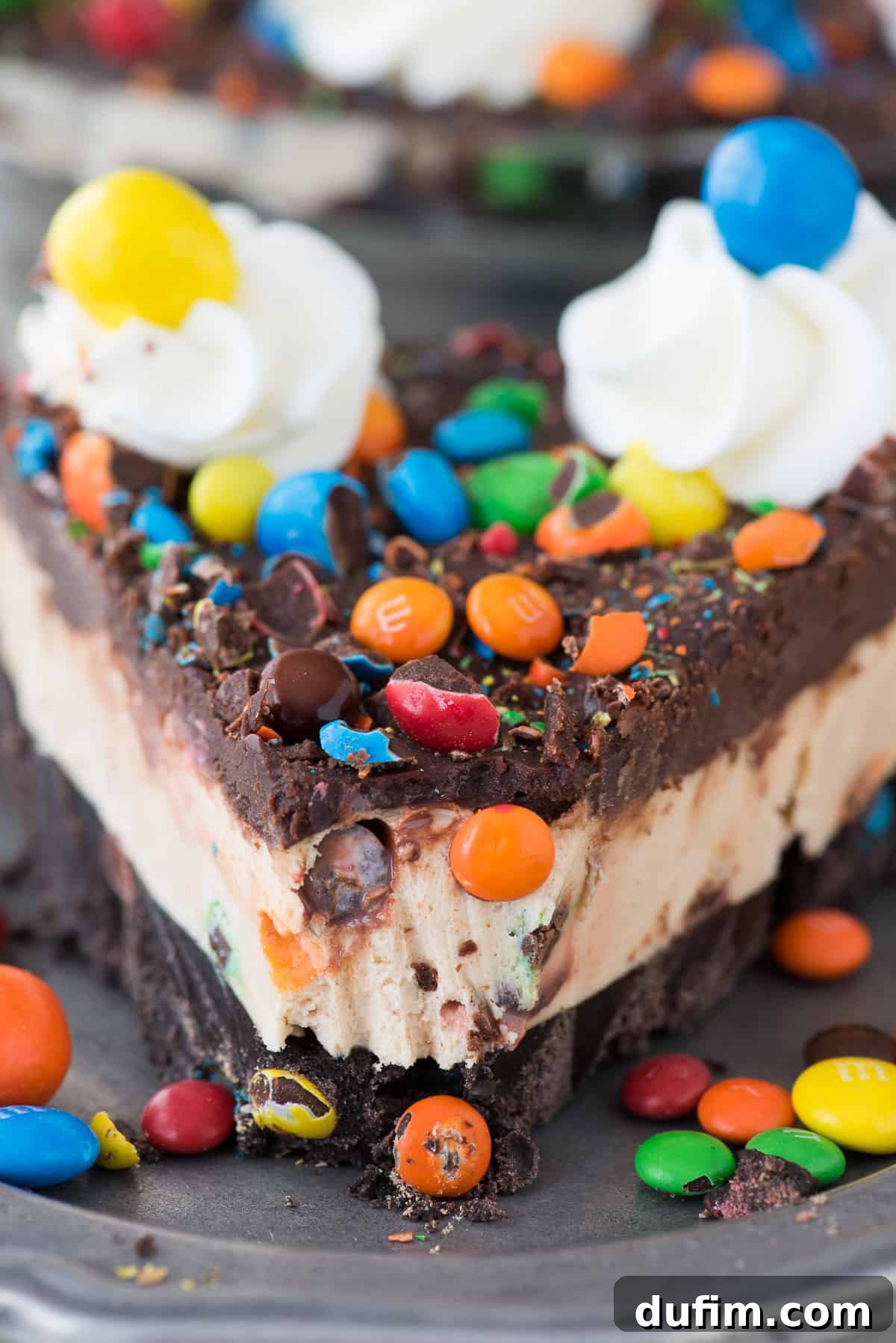 M&M No-Bake Dream Pie 4 A beautifully presented slice of M&M pie on a white plate, showcasing the distinct layers of the thick chocolate Oreo crust, creamy peanut butter filling, and luscious chocolate ganache, topped with M&Ms and whipped cream, inviting indulgence.