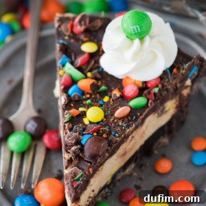 A vibrant slice of No-Bake M&M Pie on a gray plate, featuring a thick Oreo crust, creamy peanut butter filling, and chocolate ganache, generously topped with colorful M&M candies and a swirl of whipped cream. A fork rests beside the slice.