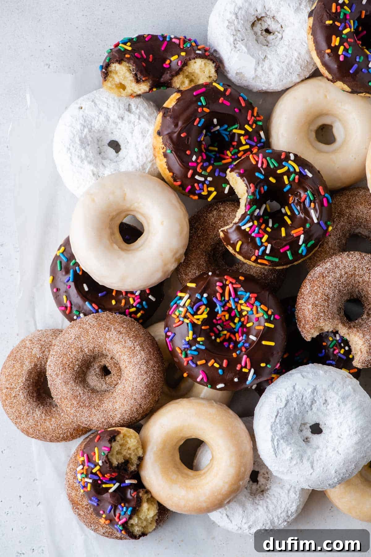 Baked donuts with a variety of glazes arranged on a clean white background, showcasing chocolate, powdered sugar, and plain glazed options.