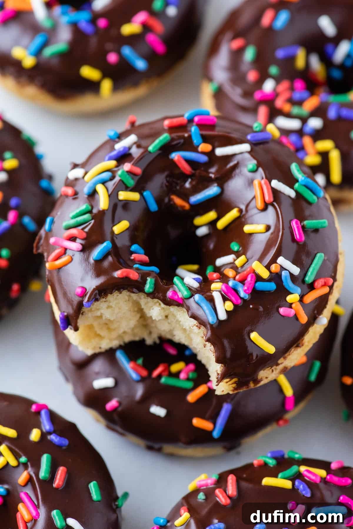A chocolate-glazed baked donut adorned with colorful rainbow sprinkles, with a bite taken out of it, sitting among other perfect donuts.