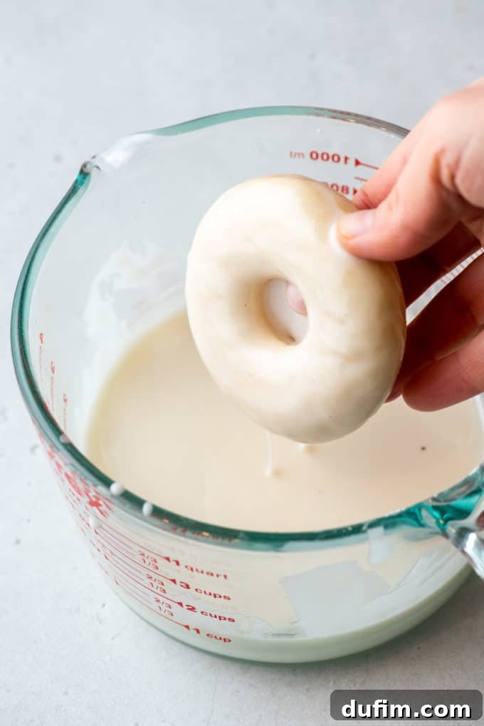 A hand gently dipping a freshly baked donut into a bowl of creamy white glaze.