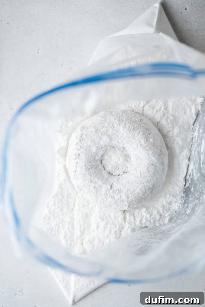 A baked donut in a Ziploc bag being coated with a generous dusting of powdered sugar.