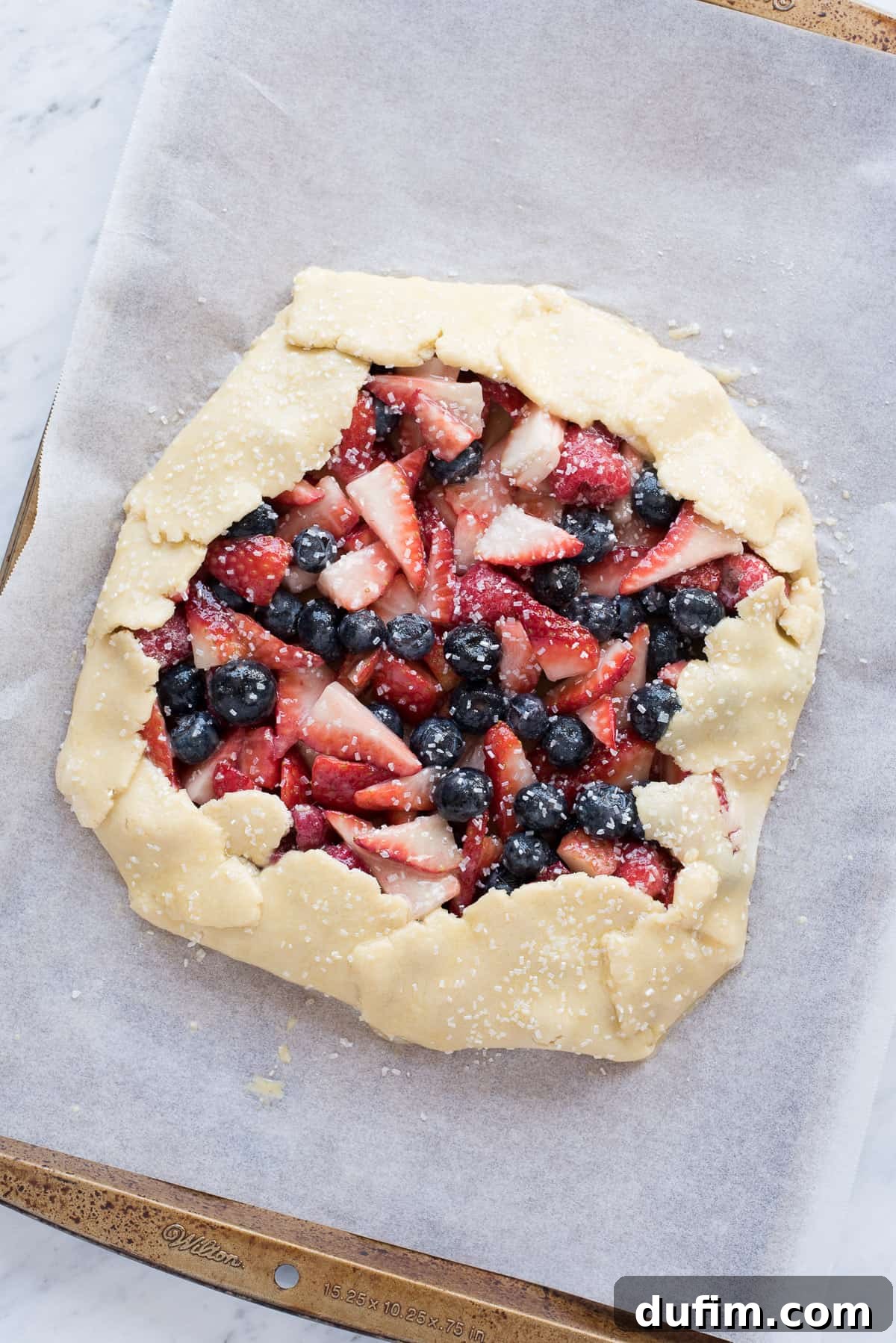 Unbaked mixed berry galette resting on parchment paper on a metal baking sheet, with the dough neatly folded around the vibrant fruit filling, ready for the oven.