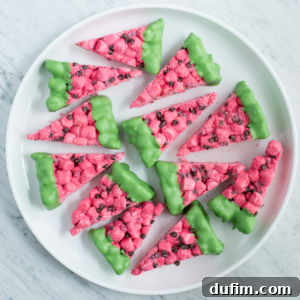 A white plate holds watermelon marshmallow slices shaped like rice crispy treats, with pink bases, chocolate chip seeds, and green-dipped edges as rinds, arranged in a circle on a marble surface.