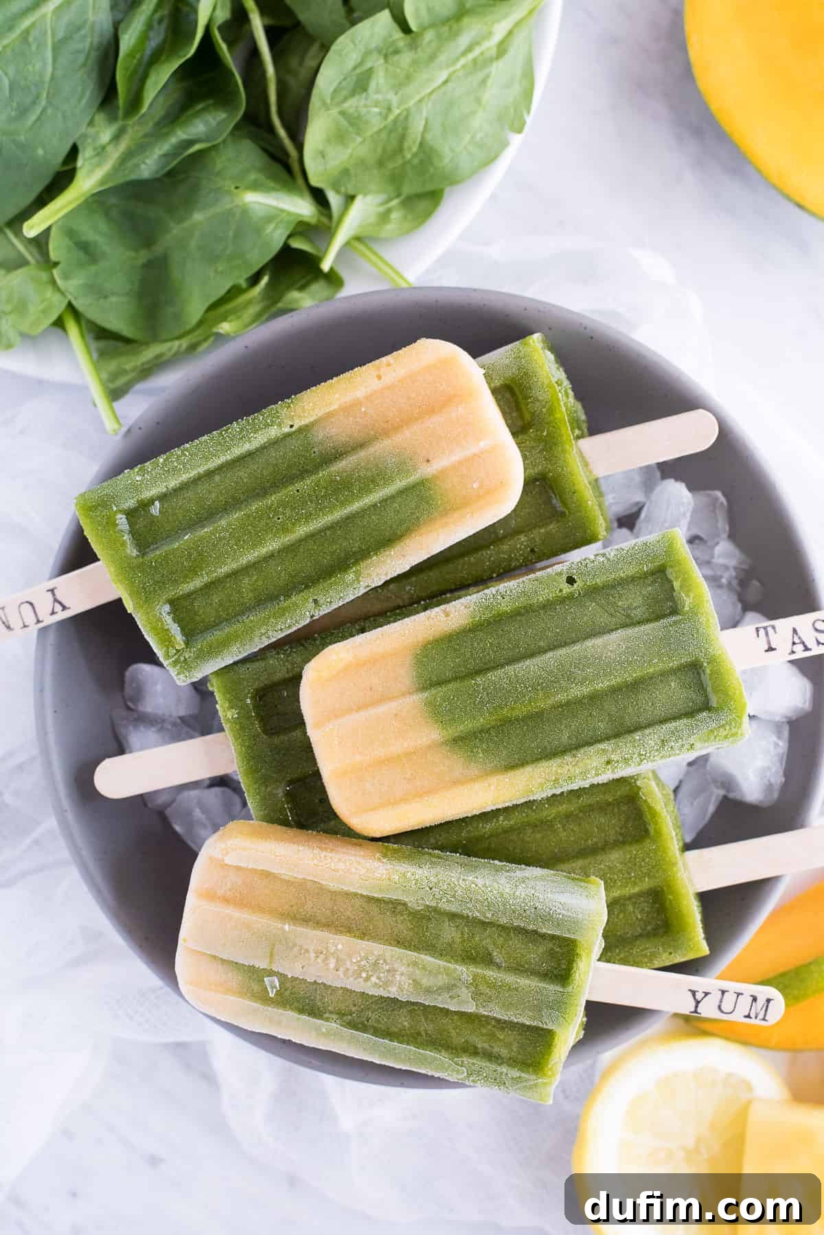 A selection of green spinach and golden mango layered popsicles standing upright in a gray bowl, against a clean white background.