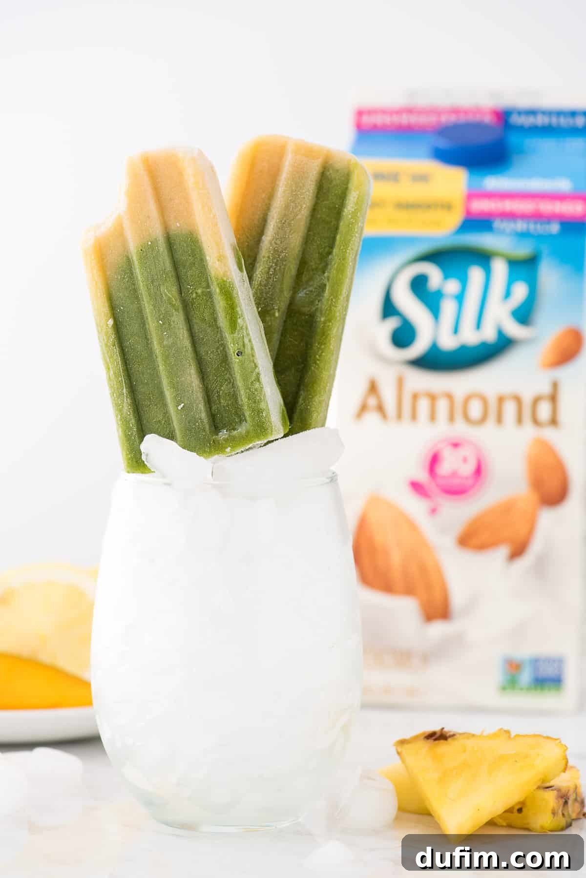 Close-up of vibrant green spinach and golden mango popsicles propped up in a glass filled with ice, with a carton of Silk Unsweetened Vanilla Almondmilk blurred in the background.