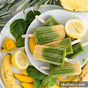 A plate filled with beautiful green machine popsicles, surrounded by fresh ingredients like spinach leaves, banana slices, mango chunks, pineapple, and lemon slices.