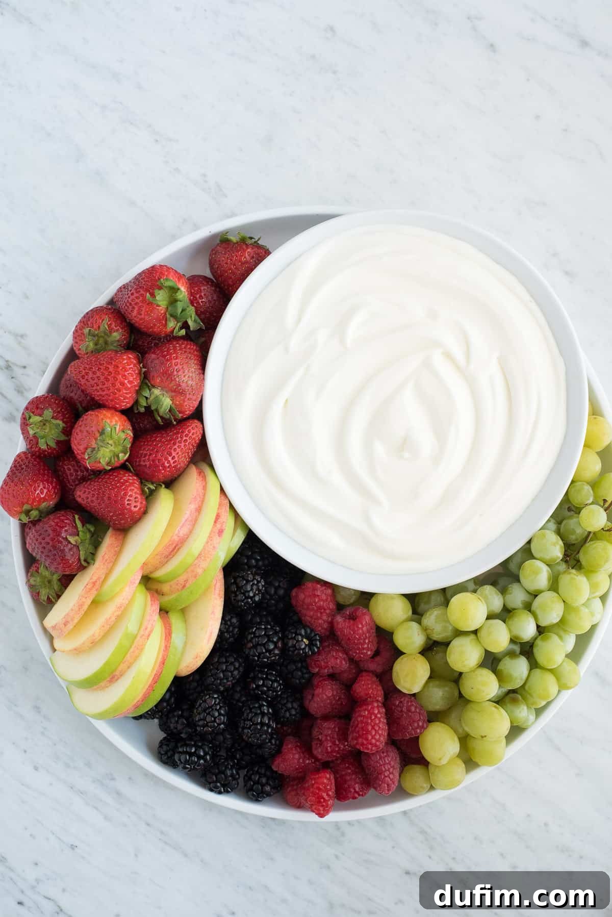 Discover the magic of this 3-ingredient fruit dip, crafted without a trace of cream cheese! An overhead view of a beautifully arranged fruit platter featuring ripe strawberries, crunchy apple slices, dark blackberries, vibrant raspberries, and green grapes, with a central bowl of creamy, white marshmallow fruit dip.