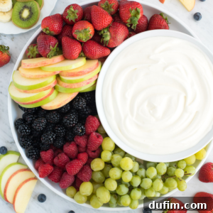 A platter with fresh strawberries, apple slices, blackberries, raspberries, green grapes, and a bowl of creamy yogurt fruit dip in the center, displayed on a marble surface.