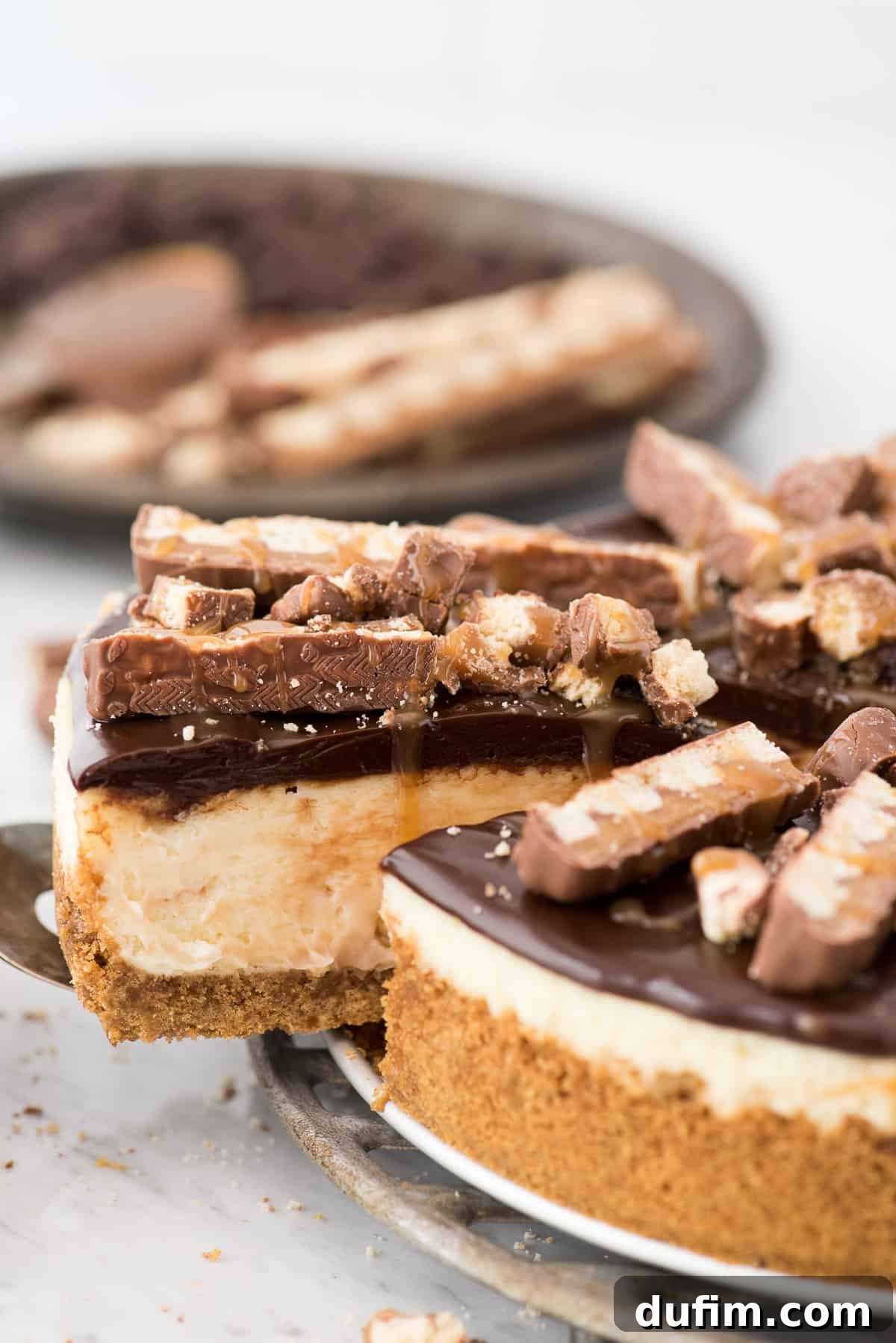 A stunning Twix Cheesecake featuring a golden graham cracker crust, a rich caramel-swirled cheesecake filling, and a generous topping of chocolate ganache, scattered with chopped Twix bars and a final caramel drizzle.
