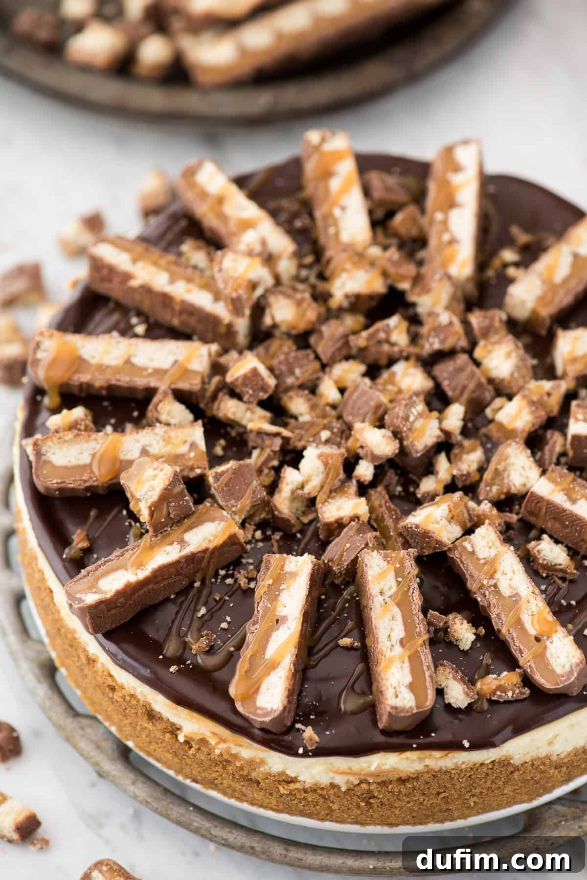 A beautifully chilled and decorated Twix Cheesecake, ready to be sliced, showcasing its perfect top with ganache, Twix pieces, and caramel drizzle.