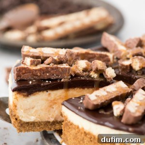 A close-up of a slice of Twix cheesecake, showing the layers of crust, caramel cheesecake, and ganache, with Twix pieces on top.