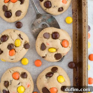 A baking tray lined with parchment paper holds several Reese's Pieces cookies topped with orange and yellow candy-coated chocolates.