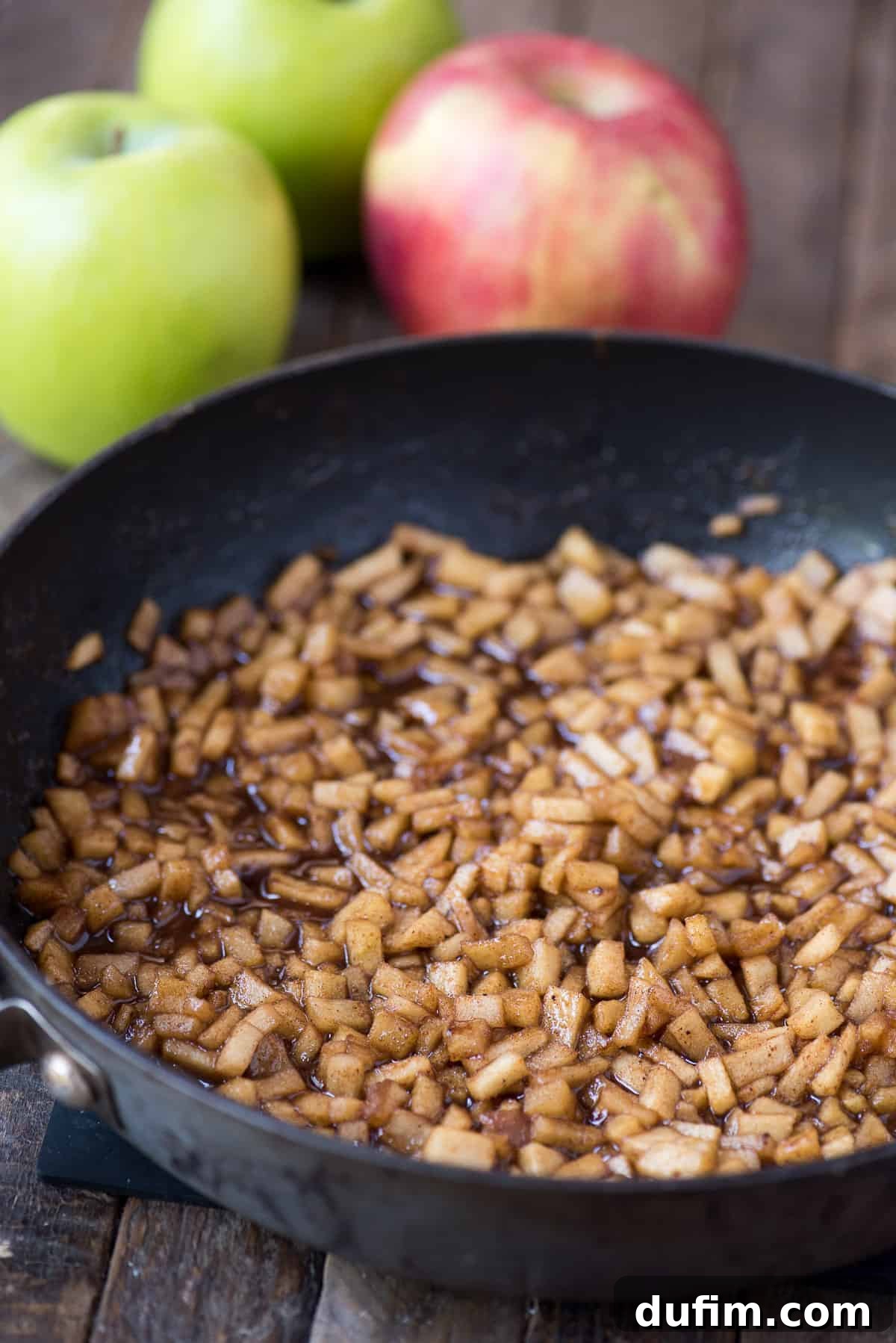 Homemade apple hand pies are a great apple dessert to make any time of the year. Watch the quick step by step video to learn how to make these mini apple pies. #applehandpies #handpies #applepie #minipie A close-up shot of caramelized apple filling simmering in a skillet pan, with fresh whole apples and cinnamon sticks arranged artfully in the background, showcasing the key ingredients.