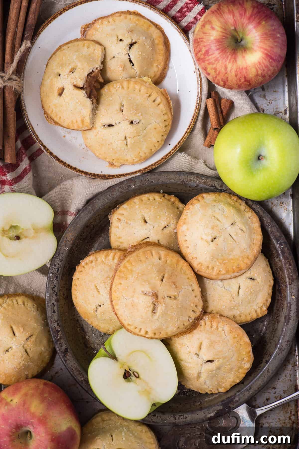 Homemade apple hand pies are a great apple dessert to make any time of the year. Watch the quick step by step video to learn how to make these mini apple pies. #applehandpies #handpies #applepie #minipie Two freshly baked apple hand pies elegantly arranged on white plates, accompanied by fresh green apples and a tan colored napkin, highlighting their golden crust and enticing presentation.
