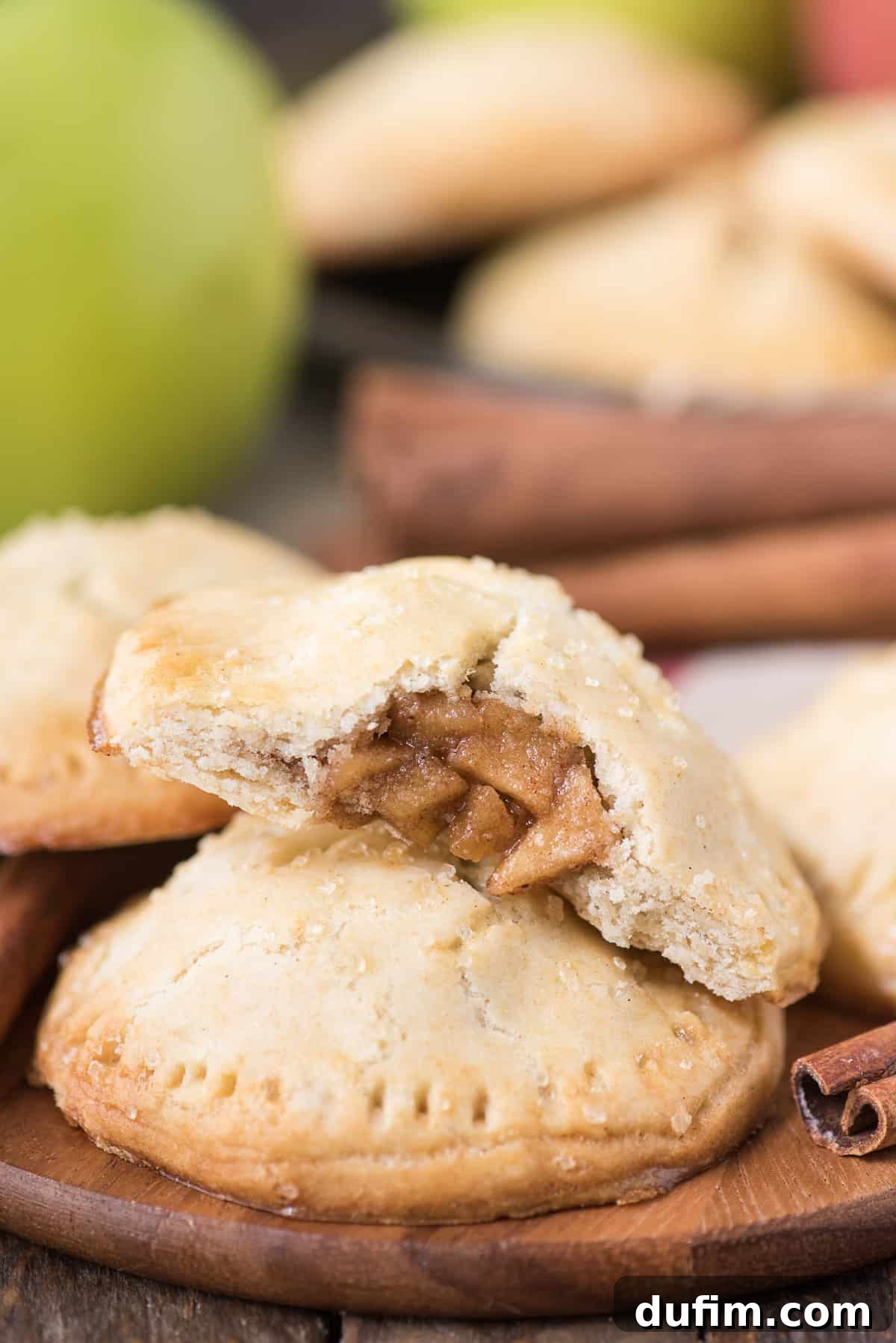 Homemade apple hand pies are a great apple dessert to make any time of the year. Watch the quick step by step video to learn how to make these mini apple pies. #applehandpies #handpies #applepie #minipie A beautifully composed shot of freshly baked apple hand pies resting on a wooden surface, with a vibrant green apple in the background, emphasizing the freshness and warmth of the dessert.
