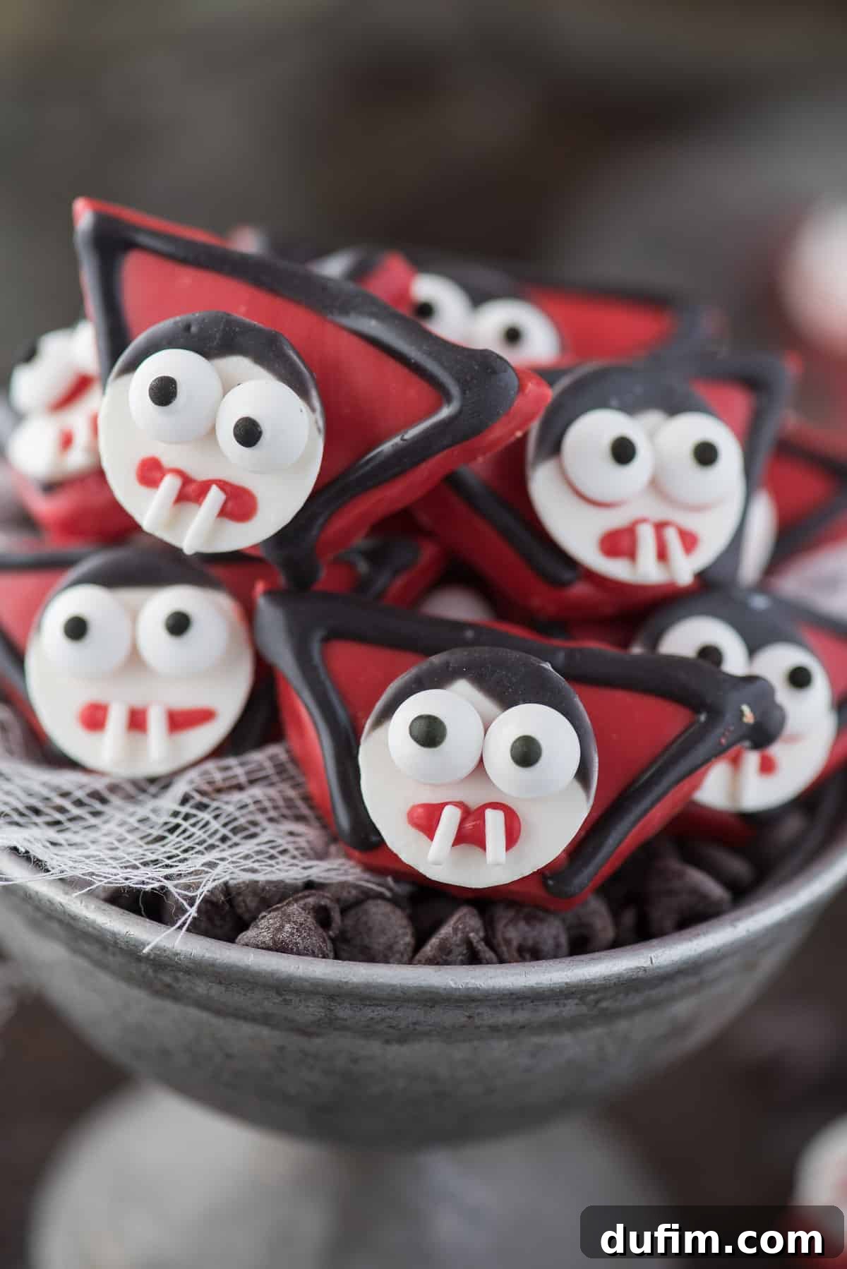 Finely detailed edible vampire treats, fashioned from graham crackers and candy melts, artfully presented in a rustic metal bowl, perfect for a themed Halloween gathering.