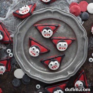 Triangular cookies decorated as vampire faces with red, black, and white icing—featuring large candy eyes and fangs—make a perfect Halloween vampire treat, arranged on a metal plate with candy pieces scattered on a dark surface.