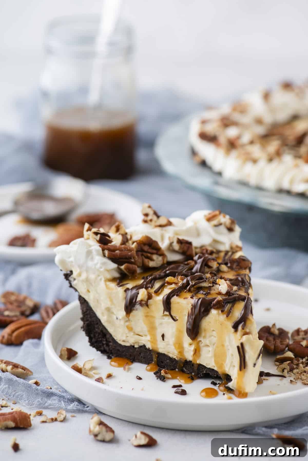 Caramel Pecan Chocolate Indulgence 5 A mouth-watering slice of Turtle Pie on a small white plate, glistening with caramel and chocolate drizzles, and topped generously with pecans. Loose pecan pieces are scattered around for an artistic touch.