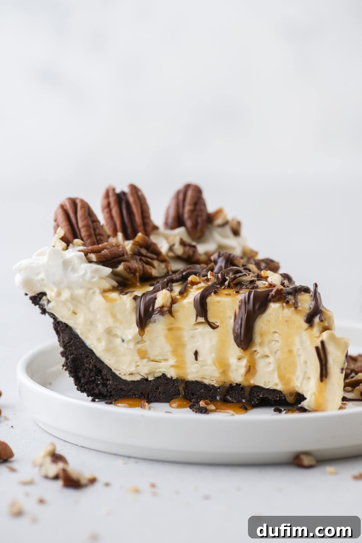 Caramel Pecan Chocolate Indulgence 9 A pristine slice of Turtle Pie on a white plate, showcasing the layered beauty of the Oreo crust, creamy filling, and the generous topping of pecans, chocolate, and caramel. The background features scattered pecans.