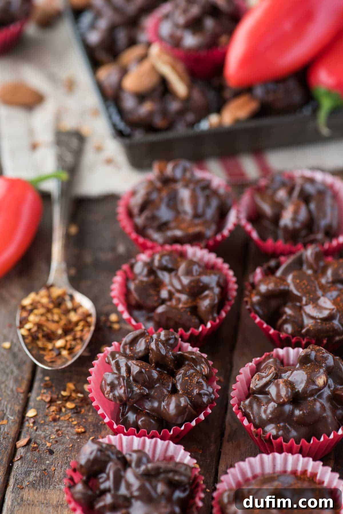 Crock pot or stove top spicy nut clusters! These dark chocolate cayenne nut clusters are popular during the holiday season. Plus there’s notes for making them less spicy or packed with heat! Glossy dark chocolate cayenne nut clusters in festive red mini muffin paper cups on a rustic wooden background, highlighting their elegant appearance.