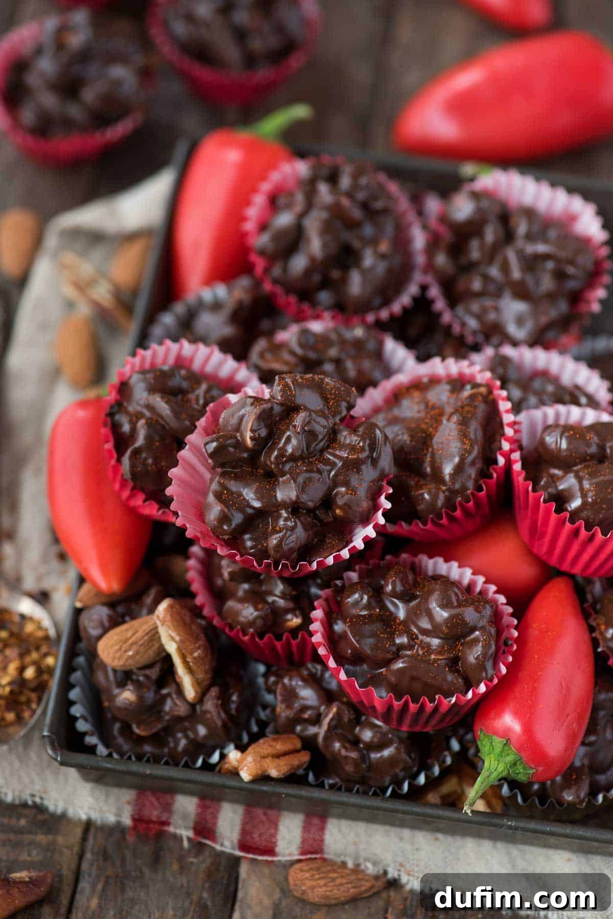 Crock pot or stove top spicy nut clusters! These dark chocolate cayenne nut clusters are popular during the holiday season. Plus there’s notes for making them less spicy or packed with heat! Another close-up of dark chocolate cayenne nut clusters in red mini muffin paper cups on a wood surface, emphasizing their perfect form and glossy finish.